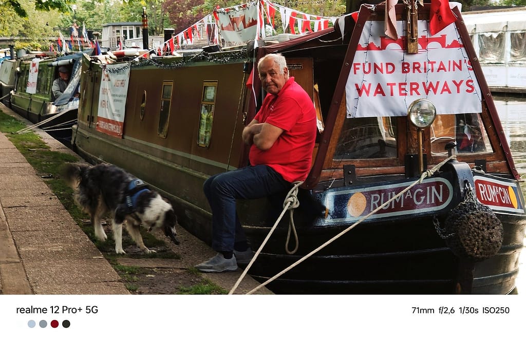 Un uomo siede fuori dalla sua barca, accanto a un manifesto che incoraggia a reinvestire nei canali, Londra, Maggio 2024